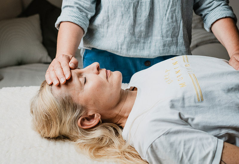 Eine Frau liegt auf einer Behandlungsliege. Die Therapeutin hat eine Hand auf die Stirn und die andere Hand auf das Hara der Klientin gelegt.